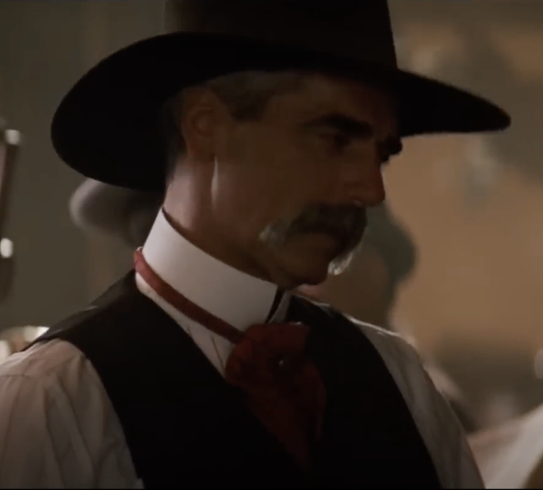 A man with a mustache is wearing a black wide-brimmed hat, a white shirt with a red bow tie, and a dark vest. He is looking to the side in a dimly lit room.