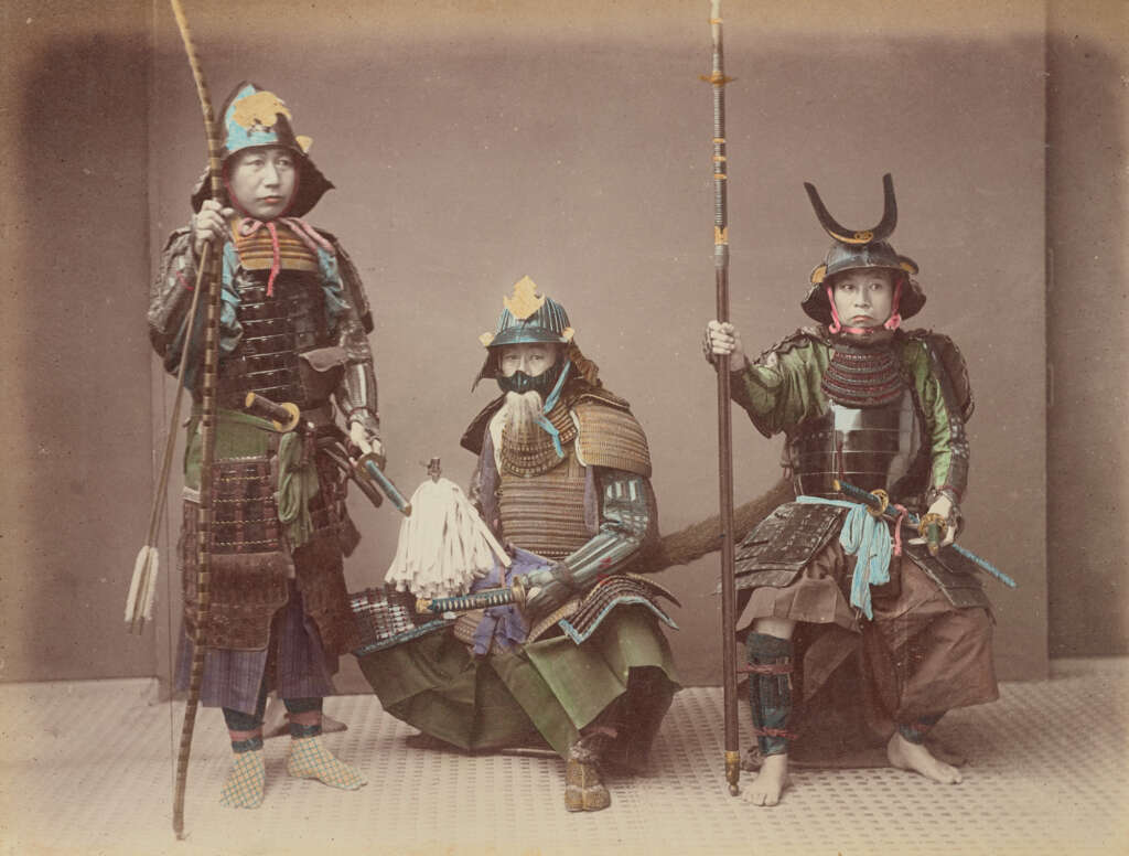 Three people in samurai armor pose against a plain background. The central figure is seated with a large sword, while the other two stand on either side, holding weapons. The armor is colorful and ornate, featuring helmets with distinct crests.