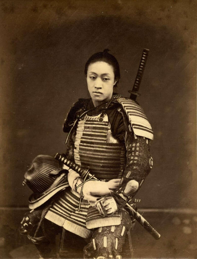 Sepia-toned photograph of a samurai in traditional armor, seated with a katana sword on their back. The armor features intricate detailing with layered plates and cords. The background is plain, highlighting the historical attire and poised demeanor.