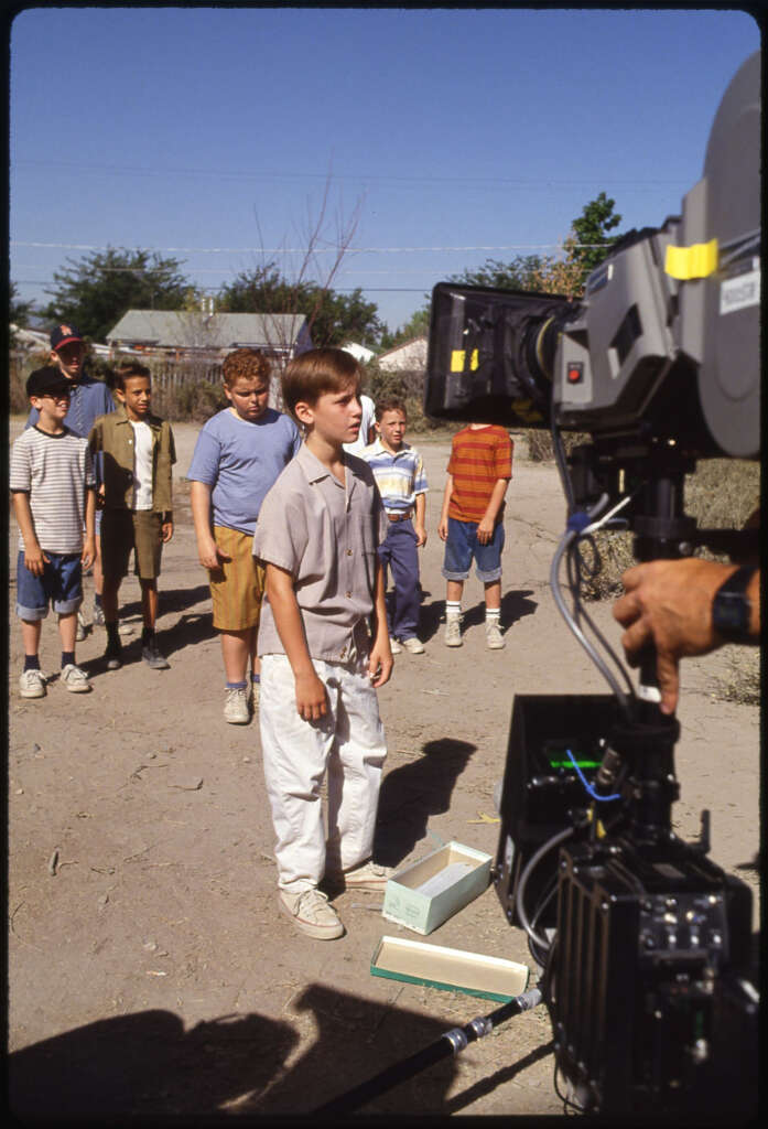 A group of boys, in casual 90s attire, stand in a dirt path facing a large film camera. The scene appears to be on a movie set, with crew members partially visible. The background shows trees and a clear sky.