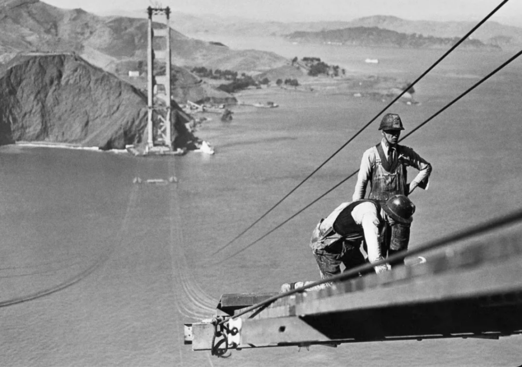 Two workers on a beam during the construction of the Golden Gate Bridge. One stands, the other kneels, both wearing safety gear. The bridge towers are visible in the background over the bay with hills in the distance.