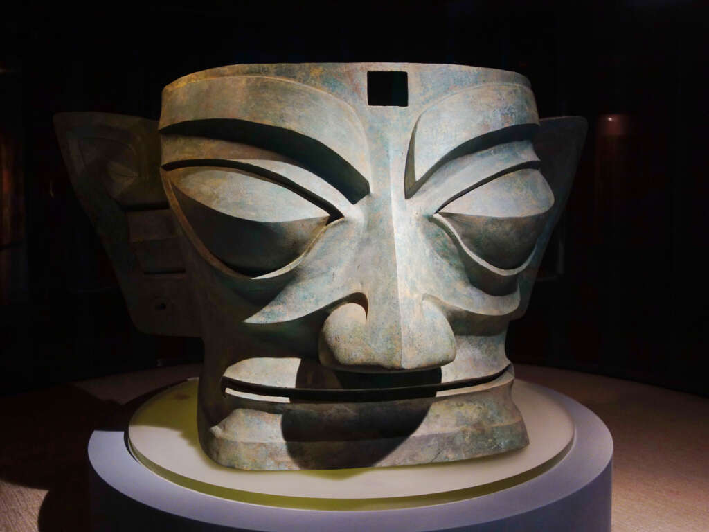 A large, ancient bronze mask with exaggerated facial features and large ears, displayed on a pedestal. The face has prominent eyebrows, slanted eyes, and a solemn expression, under a focused light in a dimly lit setting.