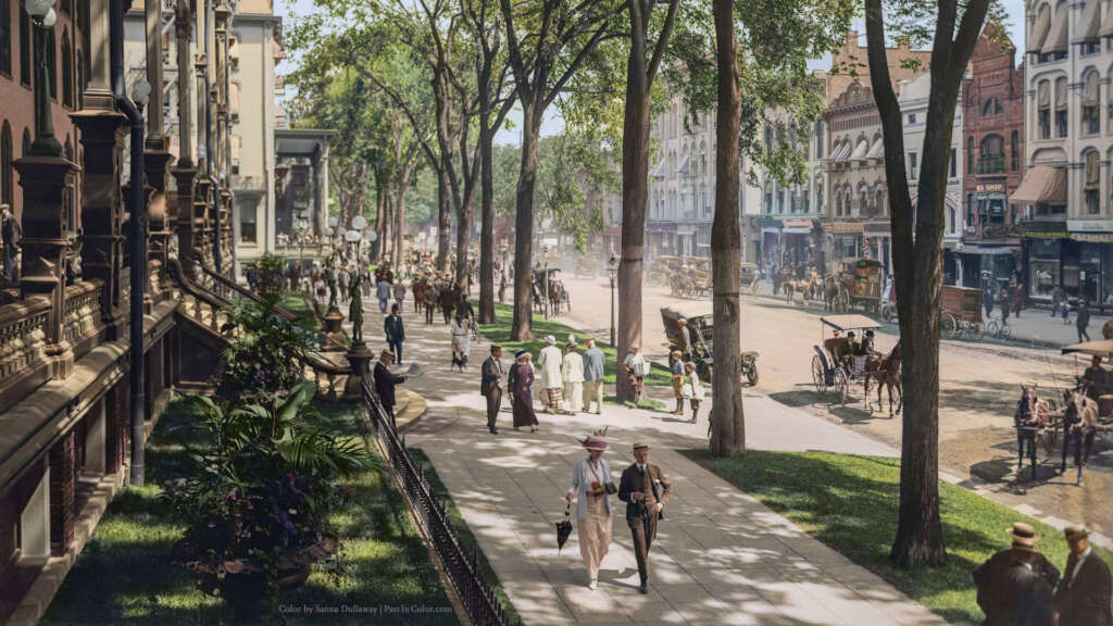 A lively street scene from the early 1900s shows people strolling along a tree-lined sidewalk, horse-drawn carriages on the road, and historic buildings with intricate facades. The atmosphere is bustling with pedestrians in period attire.