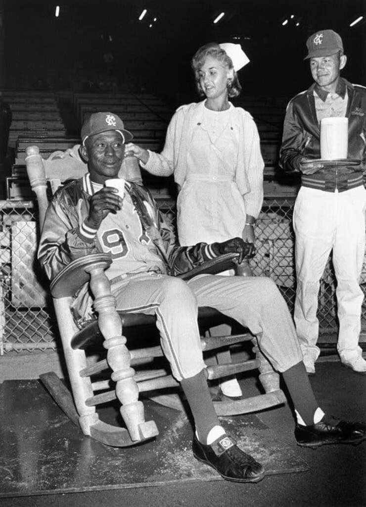 An older baseball player, wearing a "9" jersey, sits in a rocking chair on a baseball field. A woman stands beside him, and another man, holding a mug, is on the right. The background shows empty stadium seating.