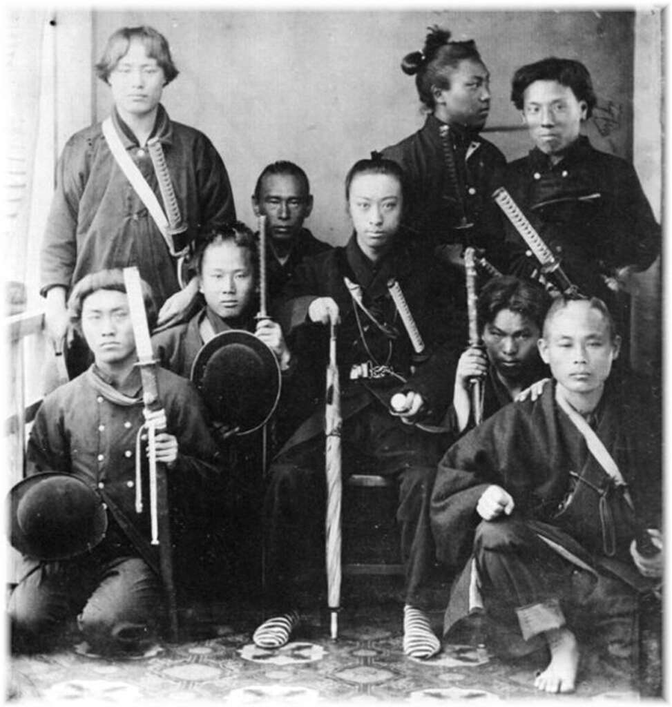 A historical black-and-white photo showing a group of nine people wearing traditional clothing. They are holding weapons such as swords and appear to be in a formal pose, facing the camera. The setting is indoors.