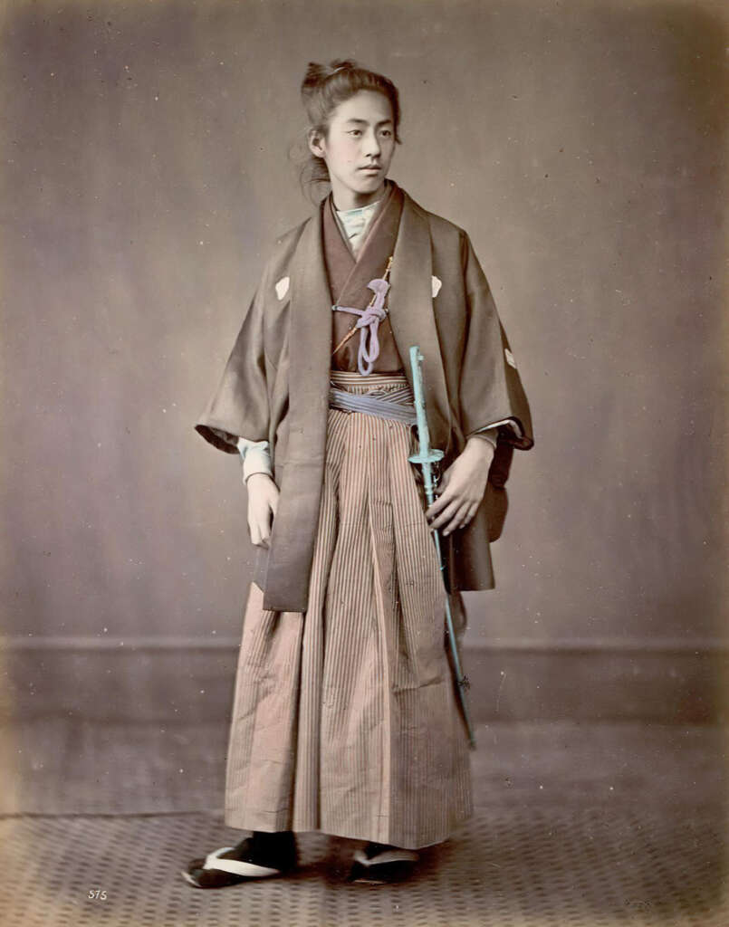 A historical photograph of a person wearing traditional Japanese attire, including a kimono and hakama, stands facing slightly to the side. They are holding a sheathed sword, with a neutral expression, set against a plain backdrop.