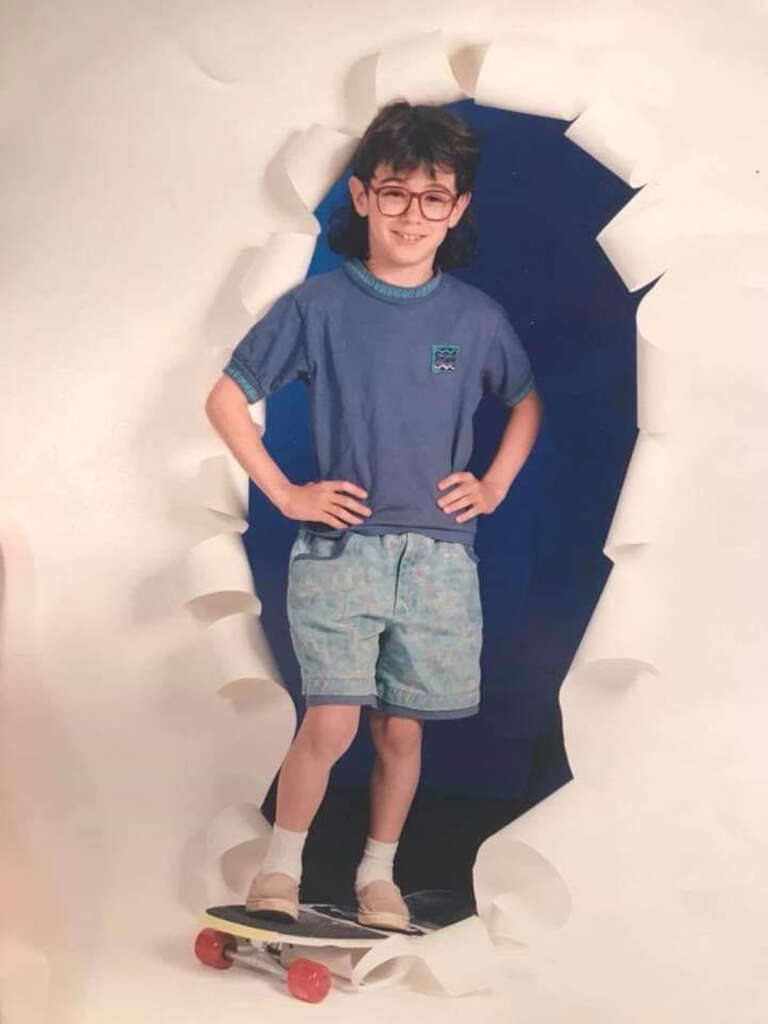 A young child stands confidently on a skateboard, posing with hands on hips. They are wearing glasses, a blue shirt, and patterned shorts. Behind them is a torn paper backdrop with a dark blue interior.