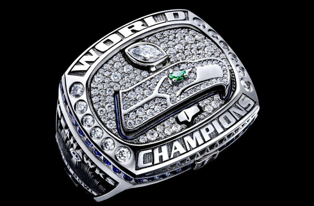 A championship ring with the word "WORLD CHAMPIONS" engraved, featuring a diamond-encrusted design with a prominent logo on top. The ring is set against a black background, highlighting its intricate details and sparkling gems.