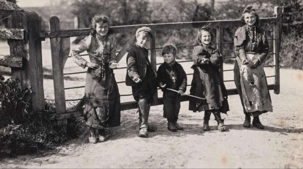 A vintage black and white photo features five children standing in front of a wooden gate. They are dressed in worn, rustic clothing. The children are smiling and holding sticks, with a backdrop of rural landscape and trees.