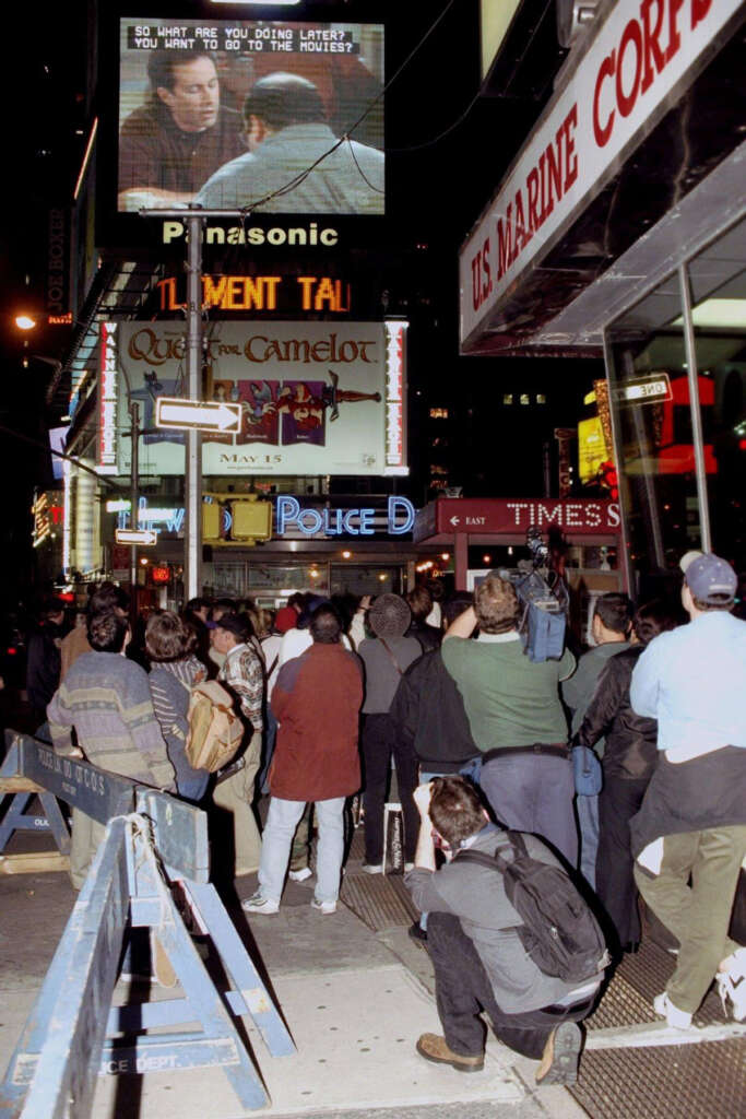 A crowd in a city street watches a large outdoor screen displaying a movie scene. Surrounding buildings have bright signage, including a Panasonic ad. It's nighttime, and the atmosphere is lively with bustling activity.