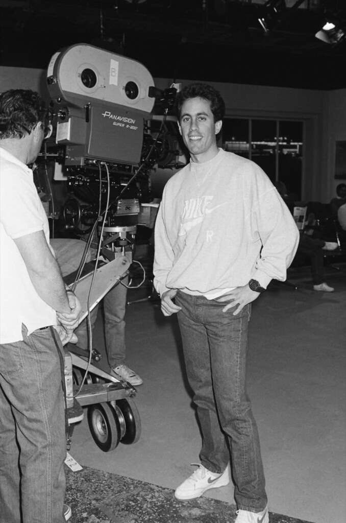 A man in casual attire stands smiling near a large film camera on a set. Another person stands in front of the camera, partially turned away. The space has a window and various equipment visible in the background. Black and white image.