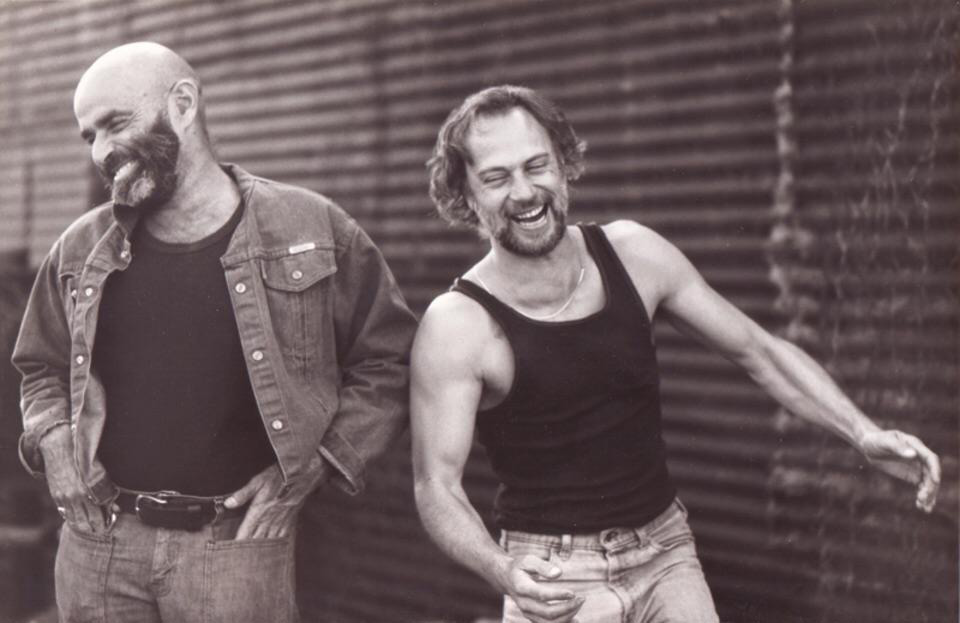 Two men laughing together in a black and white photo. The man on the left has a bald head, beard, and wears a denim jacket, while the man on the right has medium-length hair and a beard, wearing a sleeveless top. A corrugated wall is in the background.