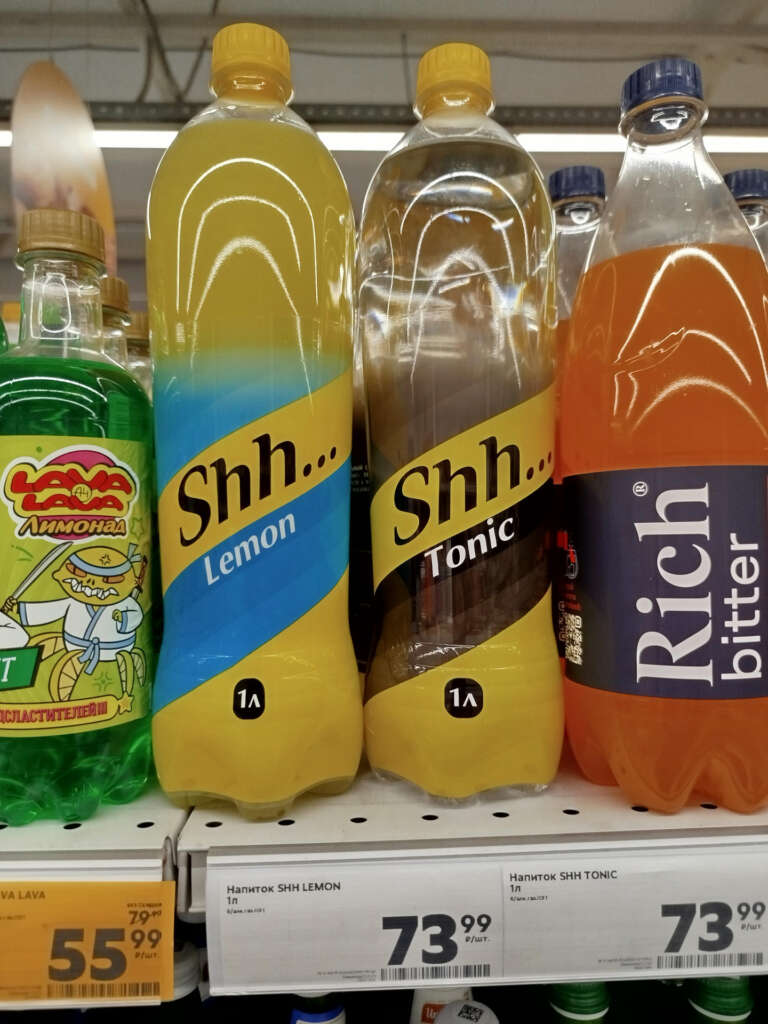 Bottles of beverages on a store shelf, including "Lava," "Shh... Lemon," "Shh... Tonic," and "Rich Bitter." Prices are displayed underneath each item. The bottles have bright and colorful labels.