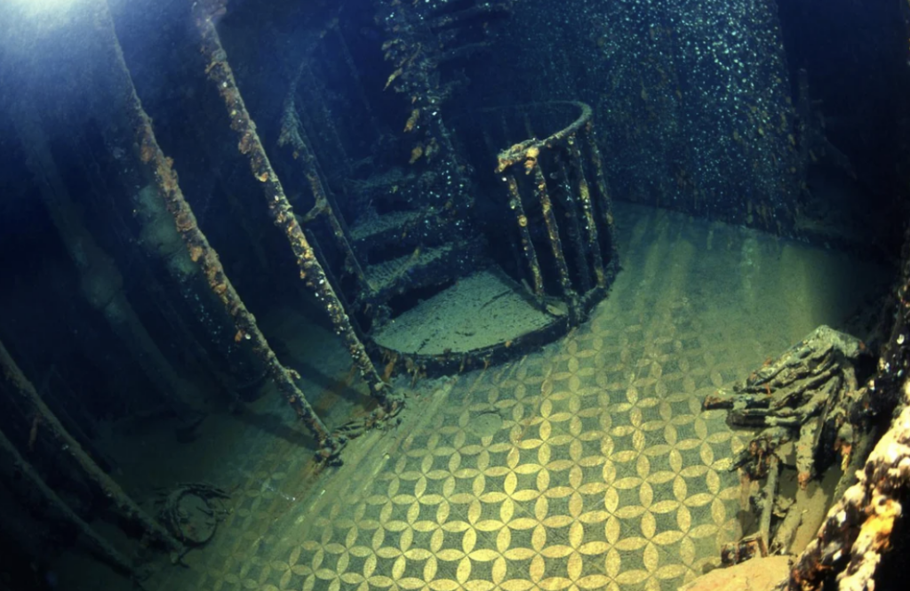 Underwater scene of a shipwreck, featuring a spiral staircase and a patterned floor. The metal structure is covered in marine growth, with dim, greenish light filtering through the water, creating a mysterious atmosphere.