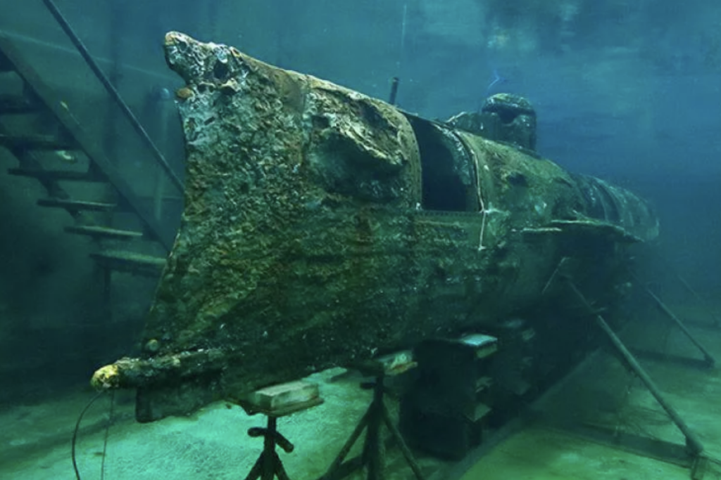 Underwater view of a corroded, historic submarine on display. It is elevated on supports, surrounded by water, giving it a mysterious and ancient appearance. Stairs are visible in the background, suggesting an exhibit setting.