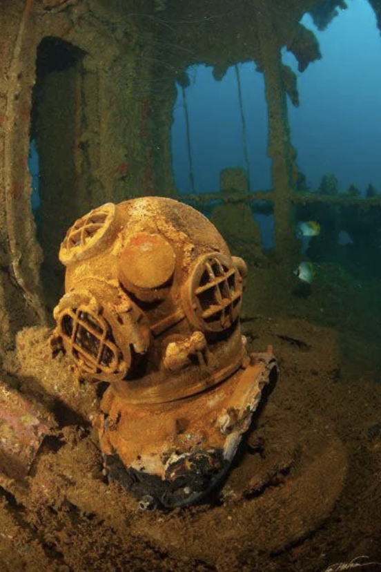 Rusty vintage diving helmet encrusted with marine growth on the floor of a sunken shipwreck. Sunlight filters through the water above, illuminating parts of the wreckage and scattered sea life.