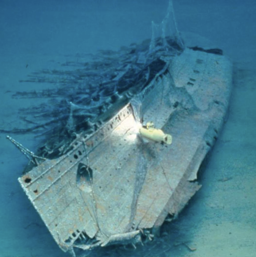 A sunken ship lies on the ocean floor, partially covered in sediment. A small submarine with a bright light is exploring the wreck. The water is deep blue, creating an eerie and mysterious atmosphere around the vessel.