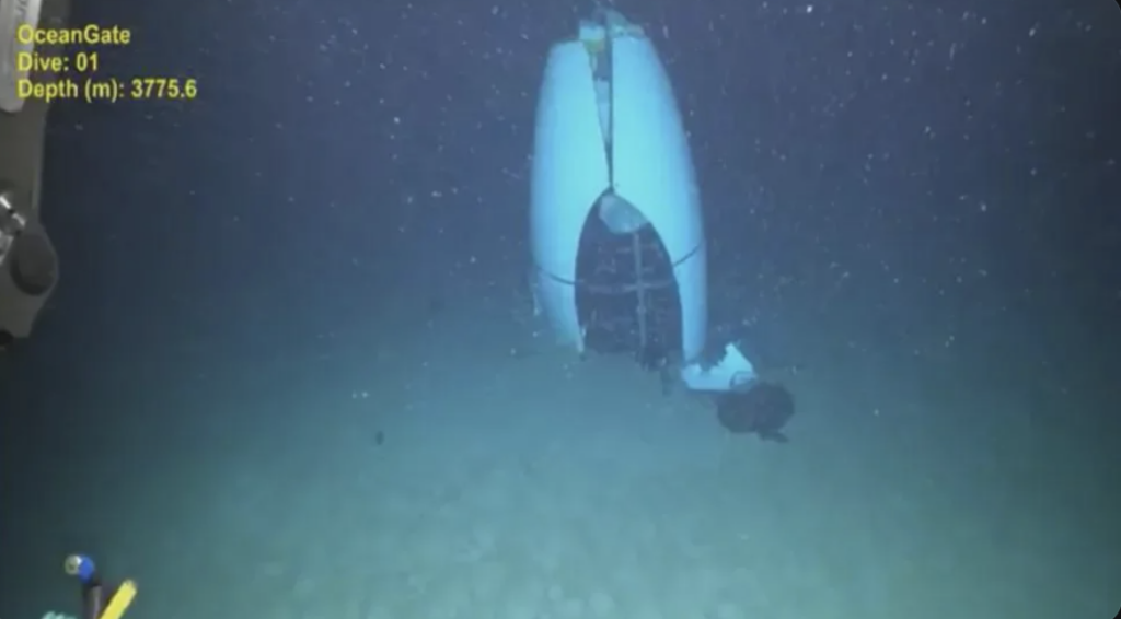 Underwater image showing a large, cylindrical object partially buried on the ocean floor. It appears to be a submersible vessel, with some marine debris scattered around. The scene is illuminated by artificial light, revealing the surrounding seafloor.