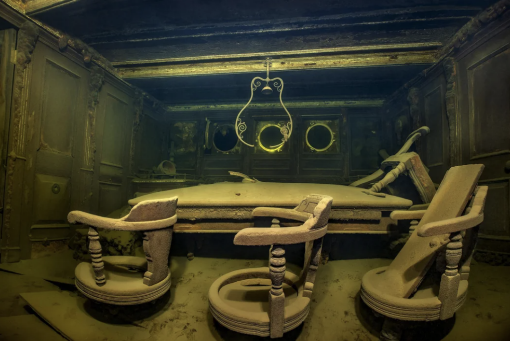 Underwater image of an old, dust-covered room with wooden chairs around a table. A decorative metal frame hangs from the ceiling, and round windows are visible on the walls, creating an eerie, submerged atmosphere.