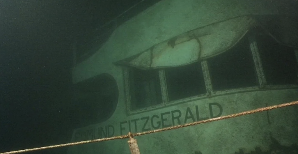 Underwater view of the shipwreck of the Edmund Fitzgerald. The ship's name is visible on a corroded and sediment-covered surface. A railing is partially visible in the foreground against the greenish underwater backdrop.