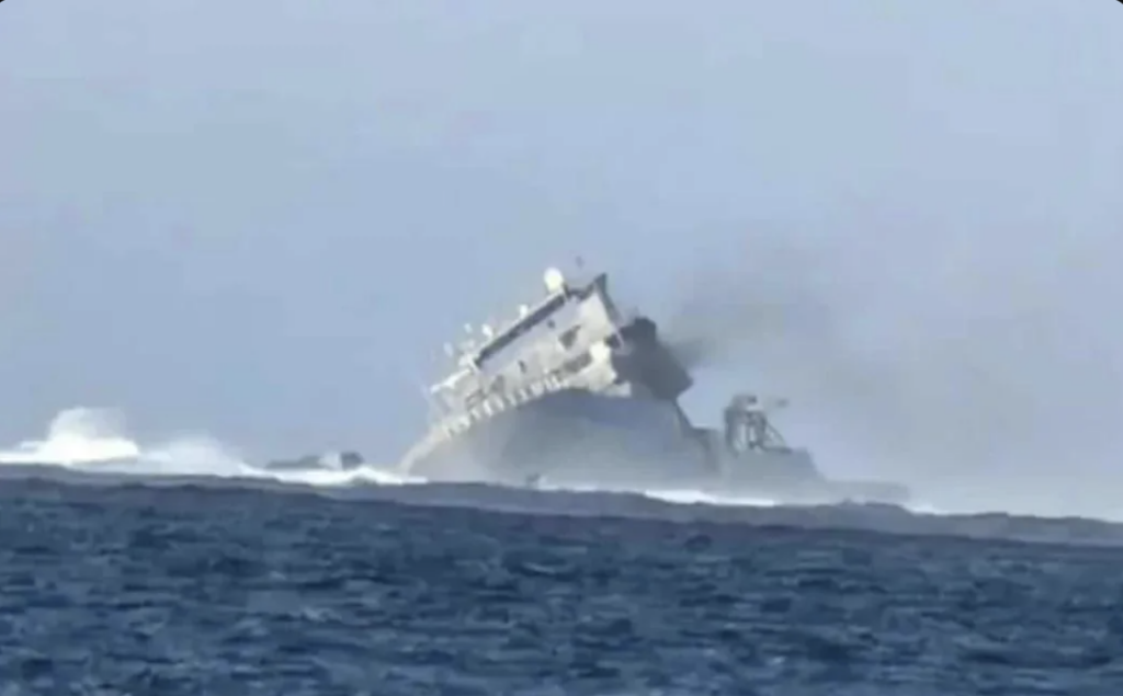 A large naval ship partially submerged and tilting to one side in rough seas, emitting dark smoke from its structure. The surrounding ocean appears turbulent, with visible waves crashing against the ship. Sky in the background is clear.