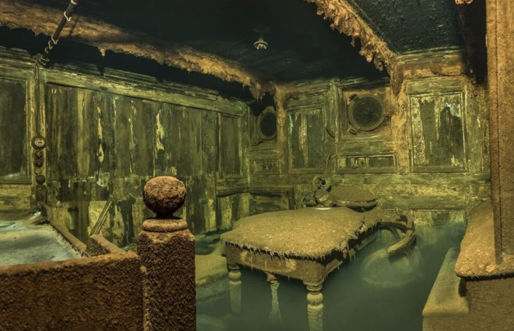 Underwater room filled with sediment and decay. A table and walls display heavy rust and deterioration. Murky water partially submerges the scene, creating an eerie, abandoned atmosphere.