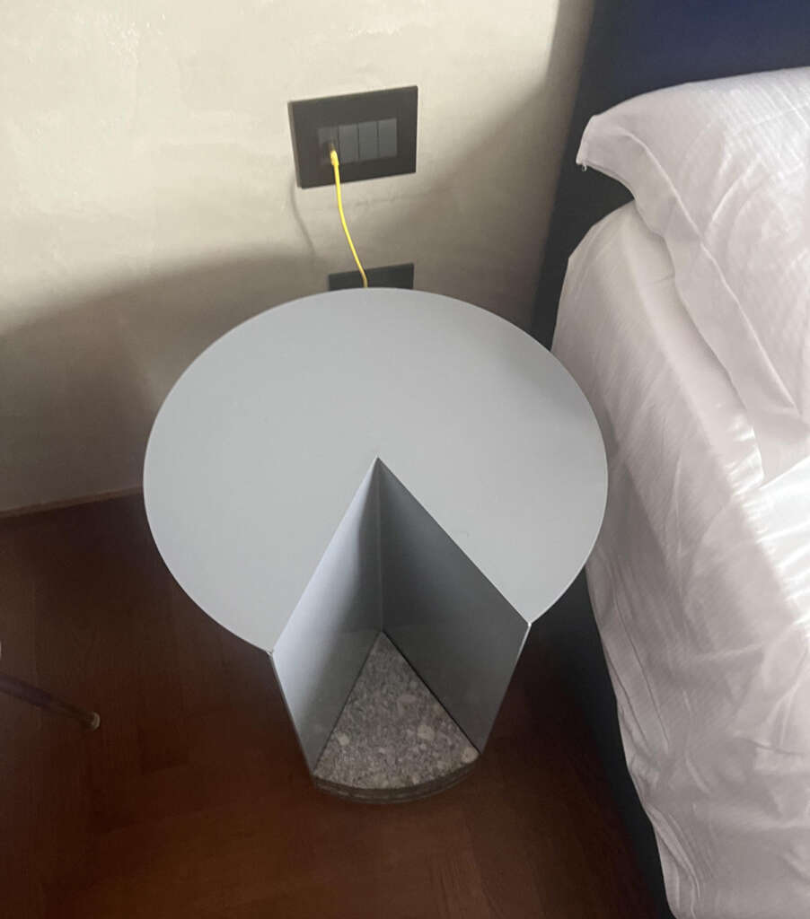 A minimalist side table shaped like a semicircle with a triangular base, placed next to a bed with white bedding. A yellow Ethernet cable is plugged into a wall socket above the table. The floor is wooden.