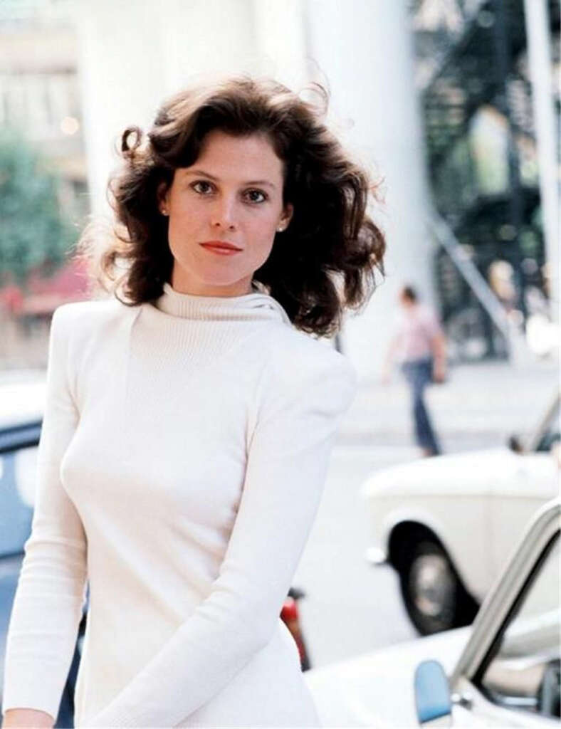 A person with wavy hair wearing a white long-sleeve top is standing outside on a street. There are cars in the background, and a person is walking in the distance.