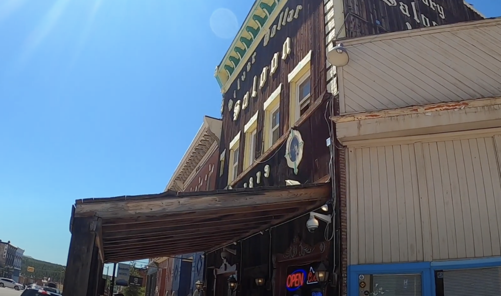 The image shows a historic downtown street with a wooden awning and colorful building exteriors. A vintage sign hangs above, along with an "Open" sign glowing near the entrance. The sky is clear and sunny.