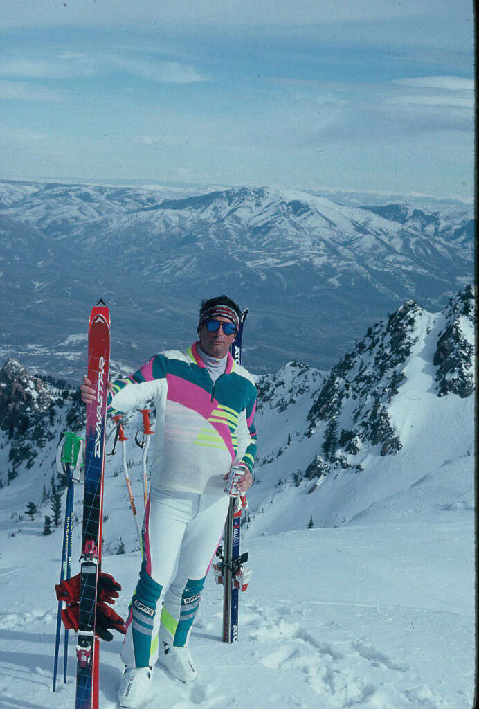 A person stands on a snowy mountain, dressed in a colorful ski outfit with sunglasses. They hold ski poles and stand beside skis stuck in the snow. In the background are snow-covered peaks and a vast mountainous landscape under a cloudy sky.