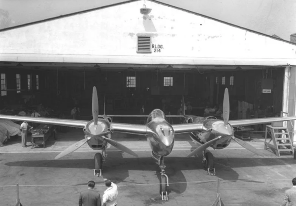 A vintage twin-engine aircraft, resembling a P-38 Lightning, is parked in front of an old hangar labeled "BLDG. 214." Several people are gathered near the plane, inspecting or working on it.