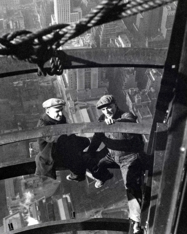 Two construction workers balance on steel beams high above a city, with buildings visible far below. They wear flat caps and heavy coats. Large cables are in the foreground, highlighting the height and precariousness of their position.