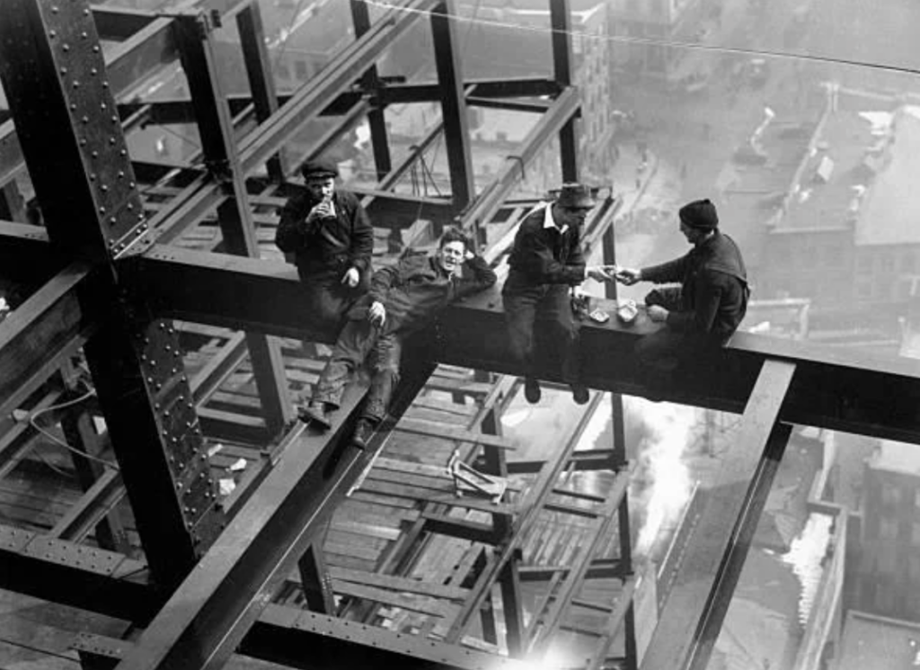 Four construction workers are sitting on a steel beam high above the city, taking a break. One is smoking, another has a beverage, and the third appears to provide food to the others. The cityscape is visible in the background below.