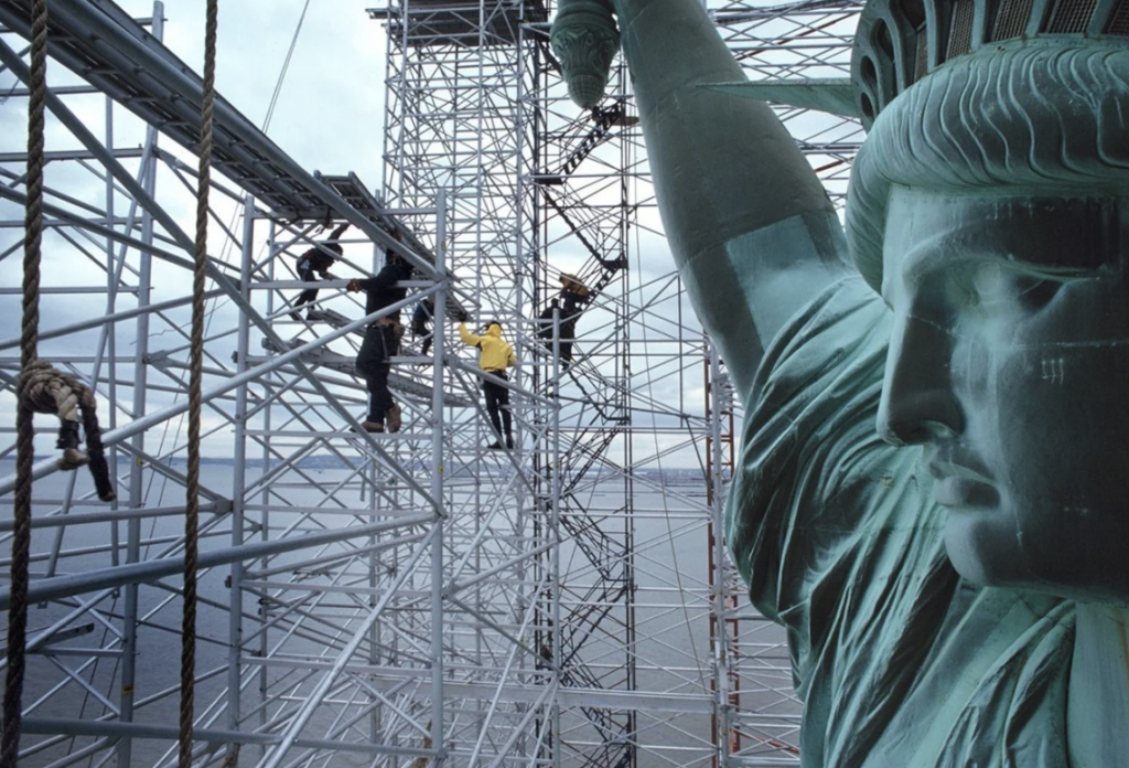 Workers on scaffolding near the head and torch of the Statue of Liberty, with water visible in the background.