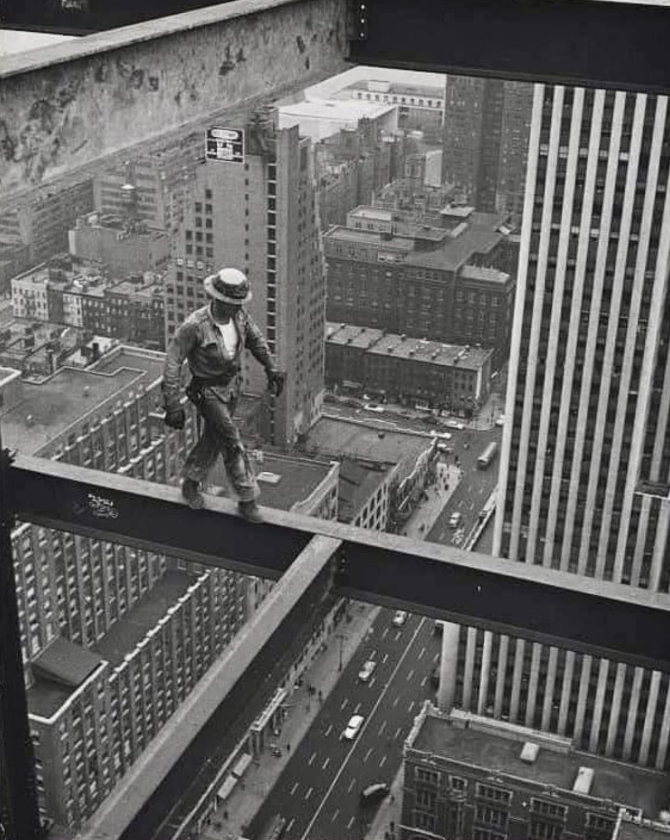 A construction worker, wearing safety gear, walks on a narrow steel beam high above a cityscape. Skyscrapers and streets with cars are visible below. The black and white photo captures a sense of danger and urban development.