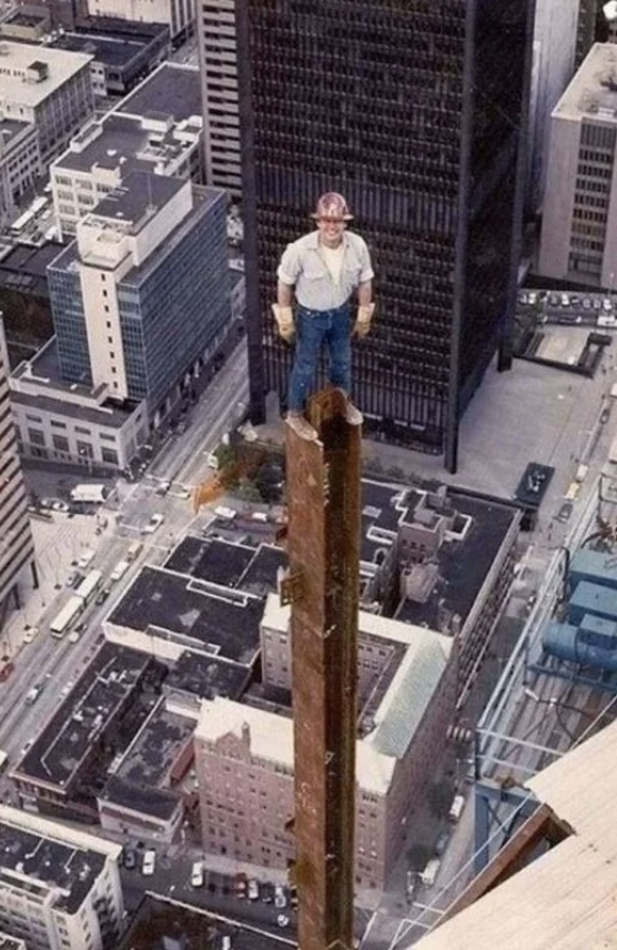 A construction worker stands confidently on top of a tall steel beam high above a cityscape, with skyscrapers and roads visible below. He wears a hard hat, gloves, and casual work attire. The image captures his elevated position and vast view.