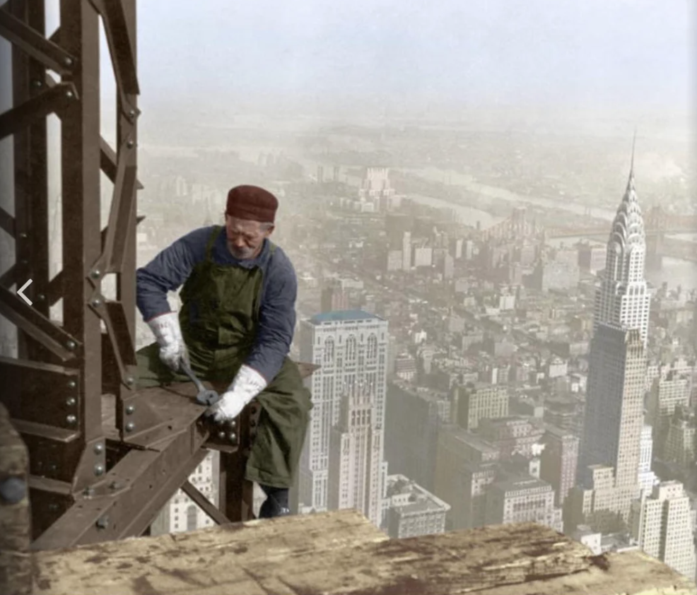 Worker in safety gear constructs a skyscraper on a steel beam high above a city skyline, with iconic Art Deco buildings visible in the background under a cloudy sky.