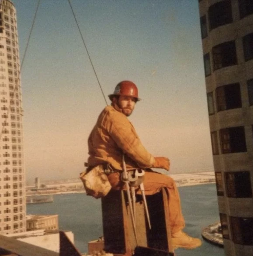 A construction worker wearing a red hard hat and brown work clothes sits on a beam high above the ground. Behind him are tall buildings and a body of water, suggesting a cityscape. The sky is clear and the scene is sunny.