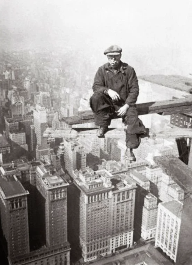 A black and white photo shows a construction worker sitting on a steel beam high above a city skyline. He is wearing a flat cap, boots, and overalls, with tall buildings visible below and a foggy sky in the background.