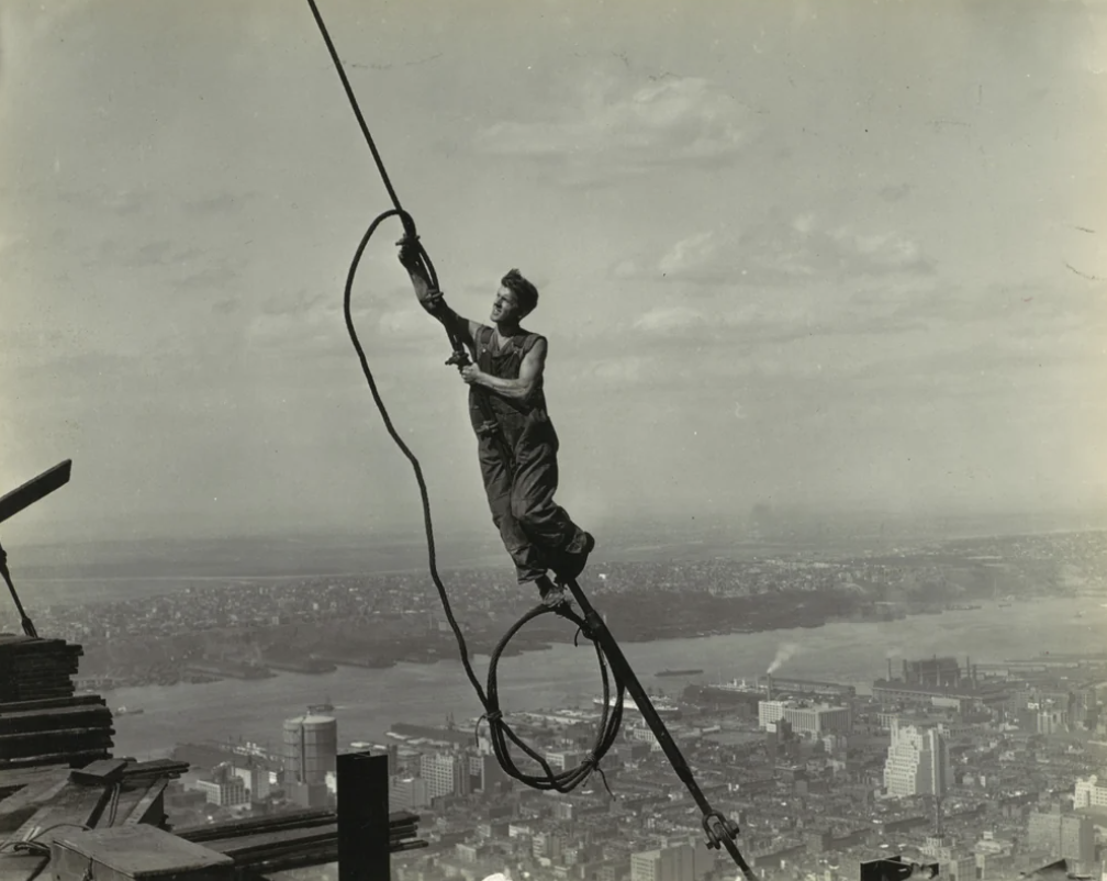 A construction worker in overalls stands on a steel beam high above a cityscape. He holds a long cable while balancing on the beam, with a vast urban landscape and river visible in the background. The sky is partially cloudy.