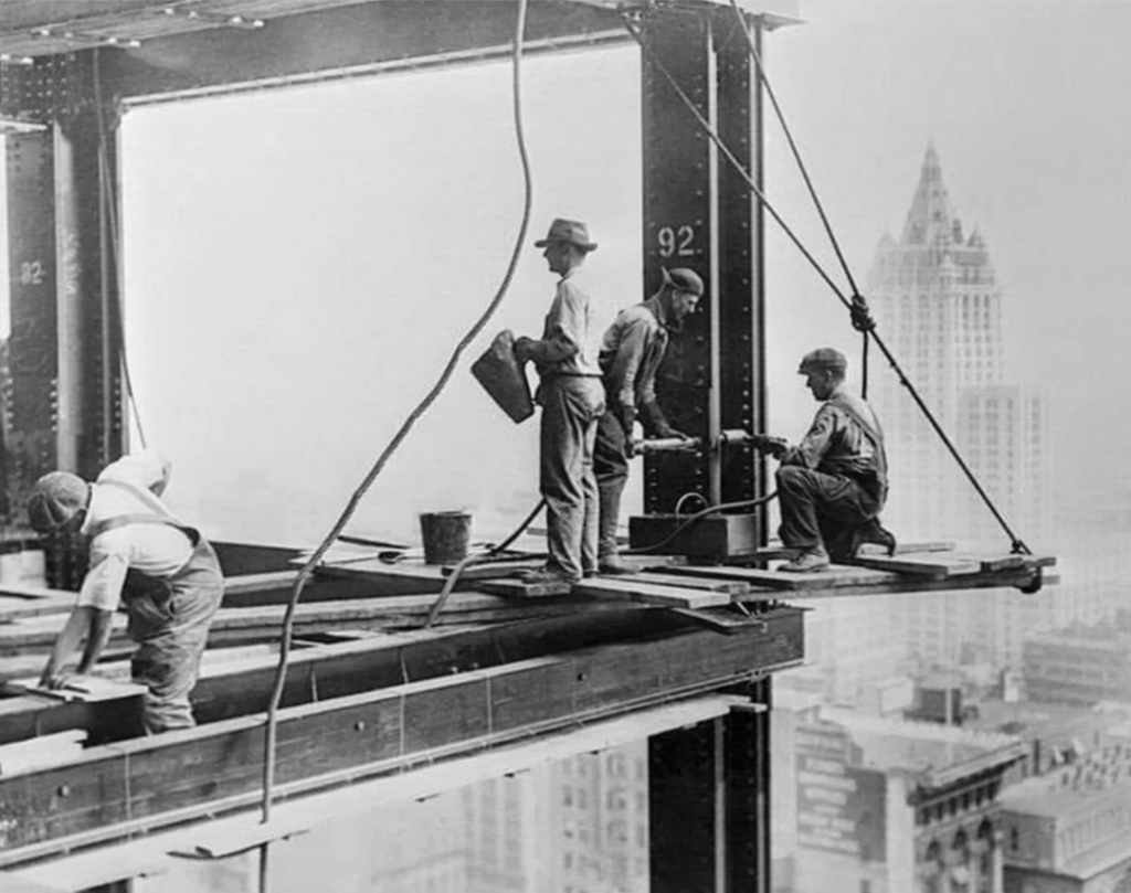 Workers on a steel beam high above a city, overlooking skyscrapers. They are engaged in construction tasks, with one holding a lunch box. The scene captures an early 20th-century construction site, highlighting the era's architecture and urban development.