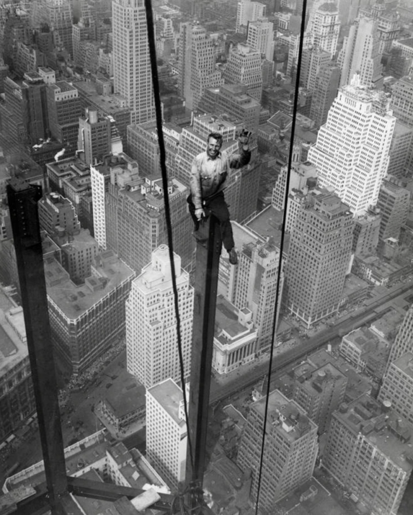 Worker balancing on a steel beam high above New York City during a skyscraper construction in the early 20th century. The bustling cityscape and iconic buildings are visible below, showcasing the vast urban landscape.