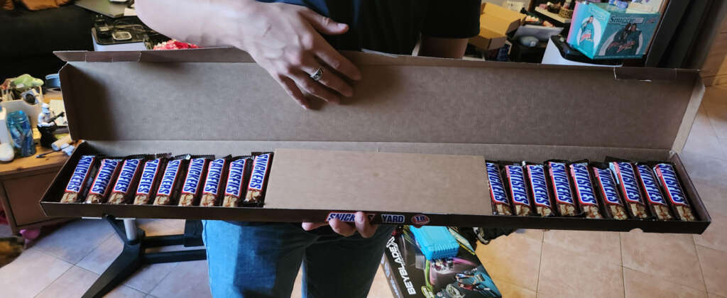 A person holding a long Snickers box containing multiple Snickers bars. The box is open, revealing rows of candy bars inside. Various household items can be seen in the background.