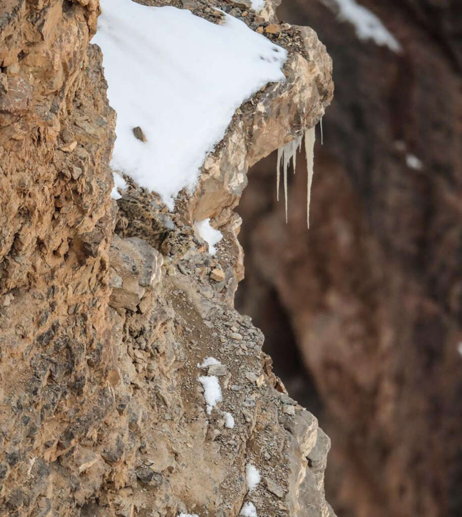 Close-up of a rocky cliff edge with patches of snow and ice. Icicles hang from the overhang, contrasting with the rugged, brown surface of the rock. Snow lightly dusts the uneven terrain.