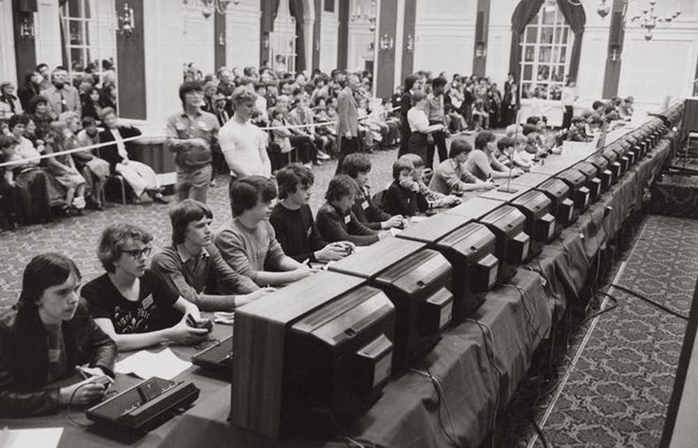 A crowded room features rows of individuals seated at desks with monitors, focused on tasks. The audience stands behind, observing the event. The setting appears formal, with ornate details visible in the background architecture.