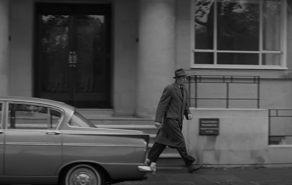 A person in a coat and hat walks briskly on a sidewalk past a parked vintage car. The background shows a building with large windows and a sign on the wall. The image is in black and white.