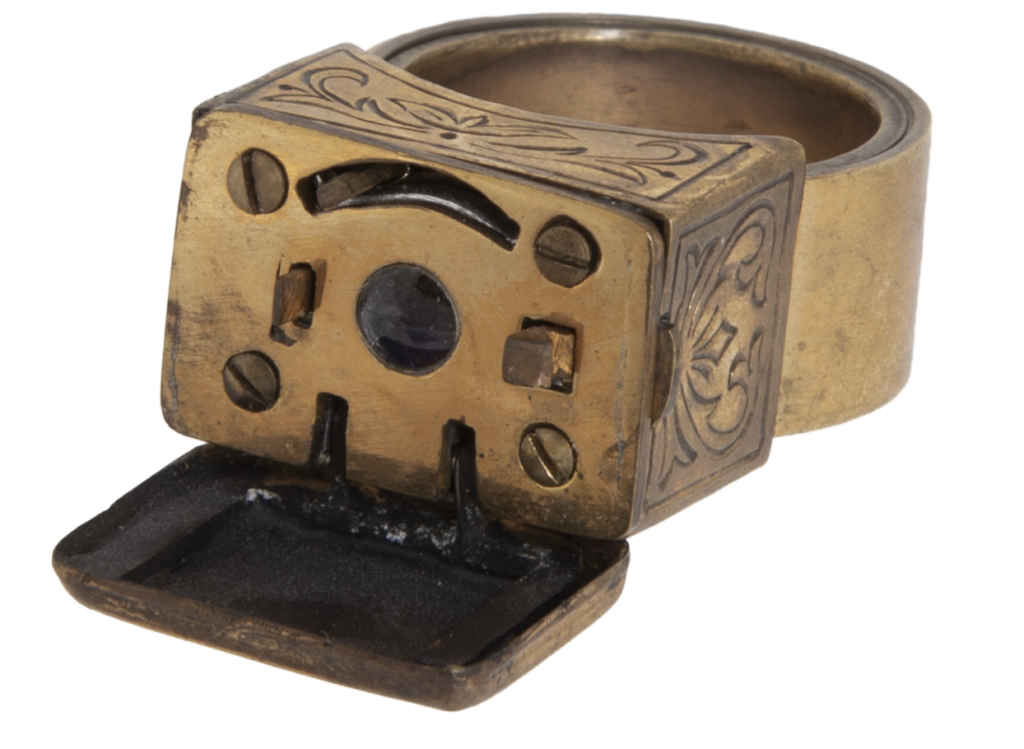 A vintage brass ring featuring an ornate design with a small hinged compartment. The compartment is open, revealing an intricate mechanism inside. The ring has decorative engravings on its surface.