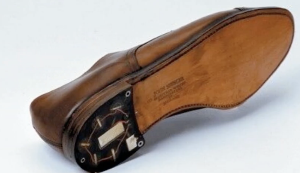 Brown leather shoe showing its underside. The sole features an integrated electronic component with visible wires, suggesting a tech-enhanced design.