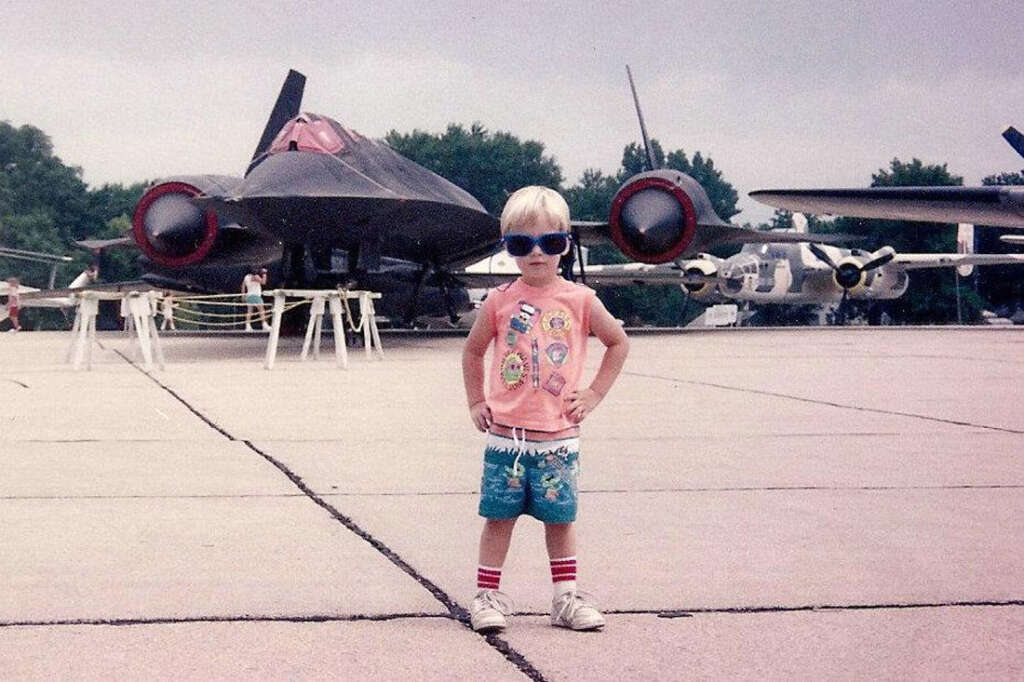 A young child with blond hair and sunglasses stands confidently on an airfield, wearing a colorful tank top, shorts, and striped socks. Behind them is a sleek, black aircraft and other planes on display under a cloudy sky.