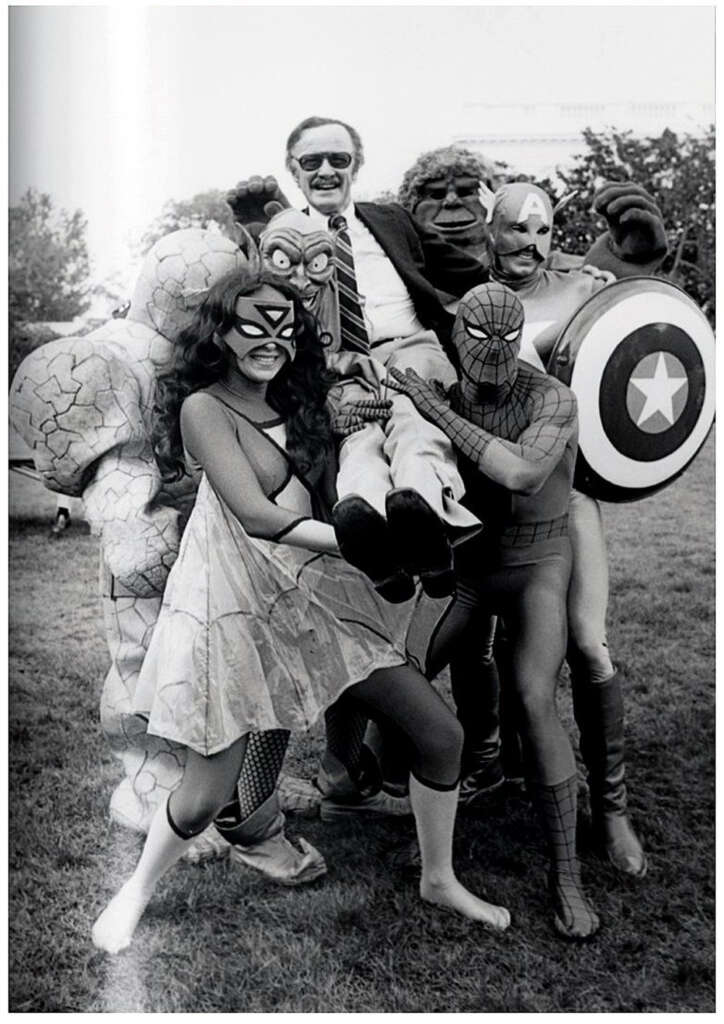 A group of people in superhero costumes, including Spider-Man, Captain America, and Hulk, cheerfully hold up a man in a suit outdoors on a grassy area. They are posing for a playful photo, smiling and embracing the moment.